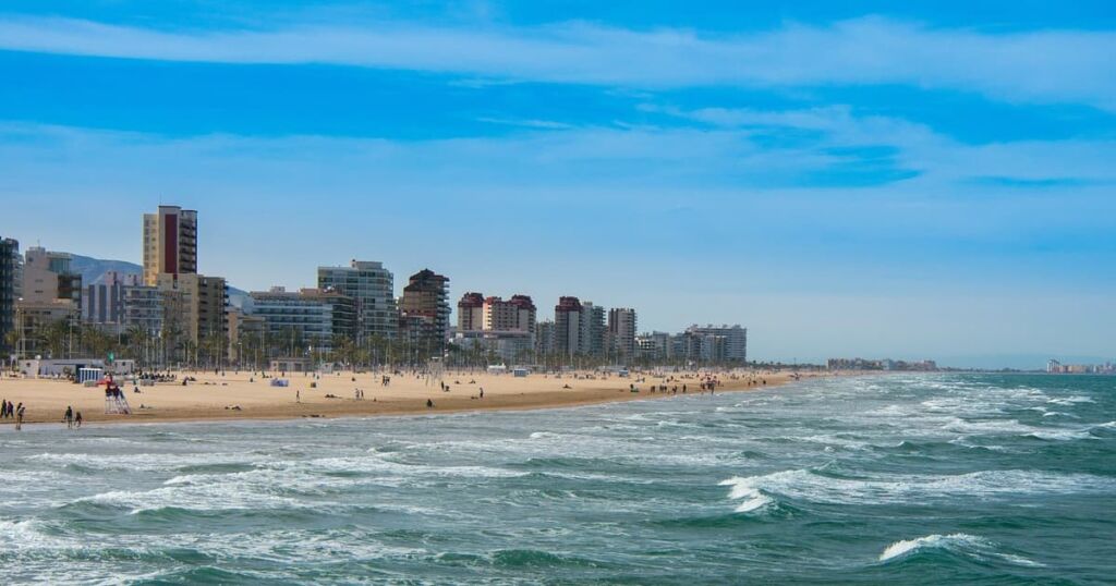 Vue de la mer agitée à Gandia, avec des vagues qui s'échouent sur la plage, la ville visible en arrière-plan, et un ciel bleu turquoise vibrant.